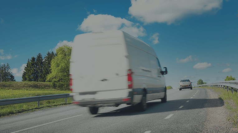 Large white van driving down a road with green grass and trees either side