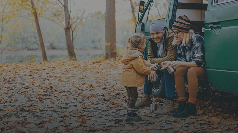 Two parents sat on the side of a green camper style van in an autumnal woodland with their young child stood in front of them