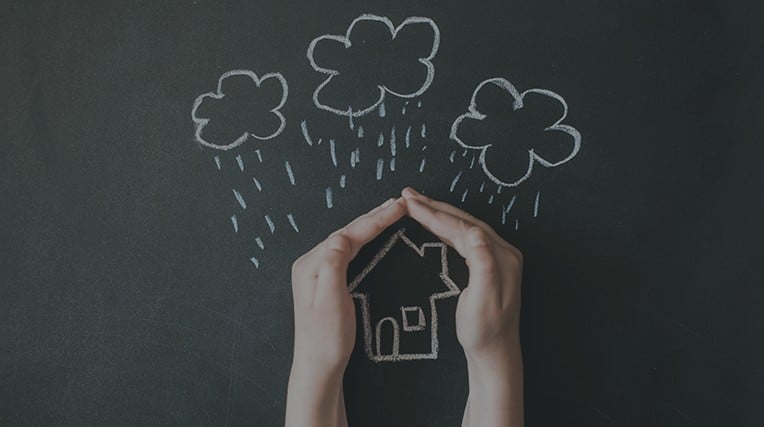 Drawing of a house with rain clouds and rain over it on a blackboard with someones hands protecting the house