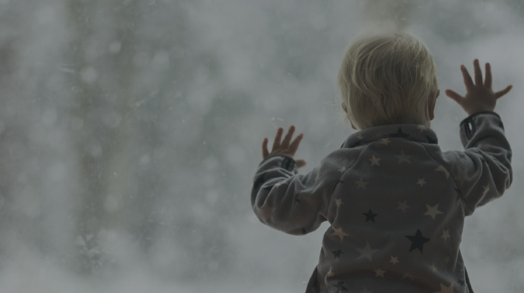 A toddler stood by a window with it's hands against the glass looking out onto a snow outside