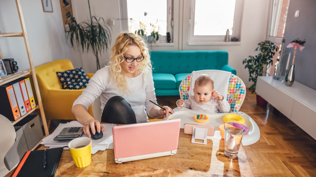 Mother and baby daughter sat at the table whilst the mother is working on a laptop